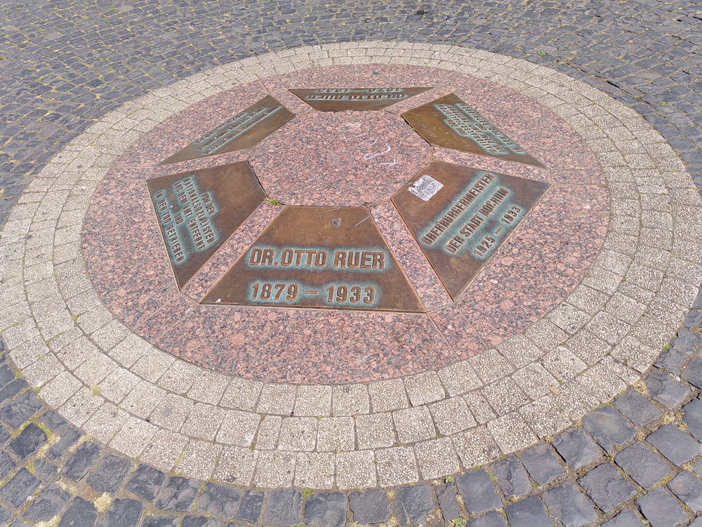 Dr. Ruer Platz Mosaikboden mit sechseckiger Form und eingelassenen Plaketten in verschiedenen Farben. Gedenktafel Dr. Ruer Platz