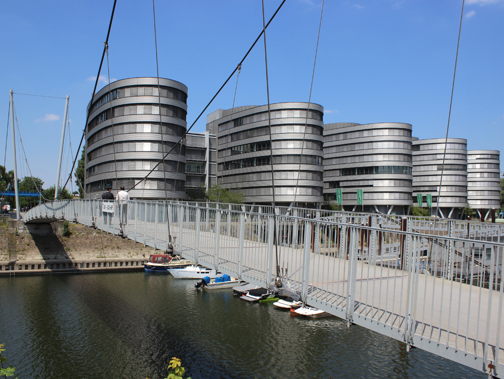 Five Boats und Buckelbrücke Moderne Gebäude mit runden, metallischen Fassaden und einer Hängebrücke über einen Kanal. Five Boats Innenhafen Duisburg