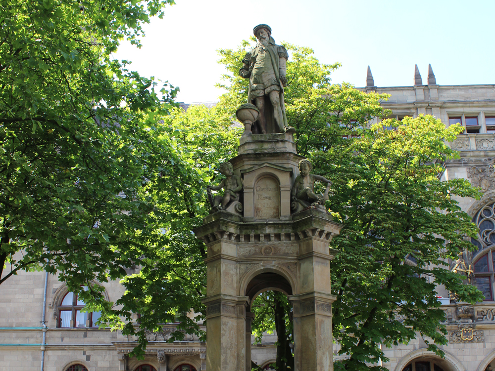 Mercatorbrunnen Rathaus Duisburg Die Statue eines Mannes steht auf einem dekorativen Sockel, umgeben von Bäumen. Mercatorbrunnen Rathaus Duisburg