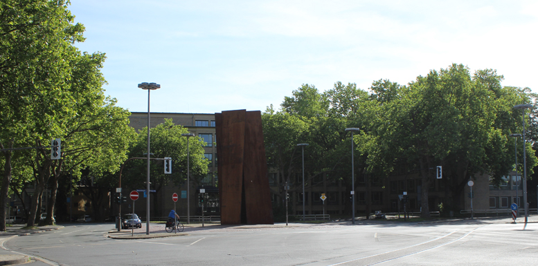 Terminal Bochum, Richard Serra Ein rustikales, metallisches Kunstwerk vor modernen Gebäuden und Bäumen in einer Stadt.
Terminal Bochum von Richard Serra