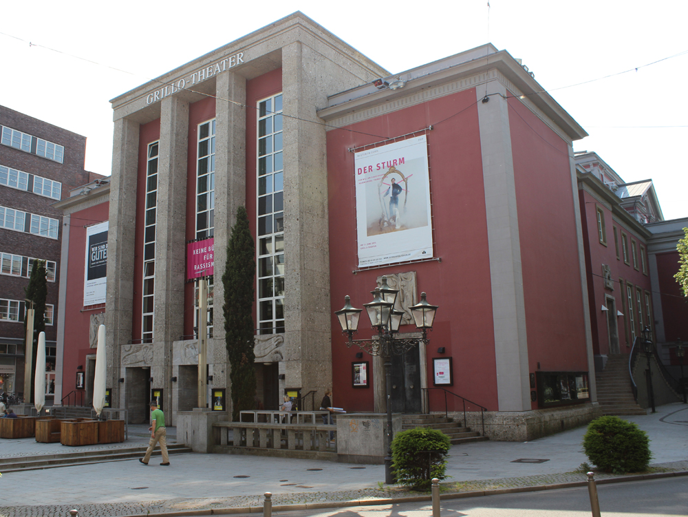 Grillo Theater Historisches Gebäude mit roter Fassade und Plakaten, umgeben von Pflanzen und Straßenlaternen. Grillo Theater Essen