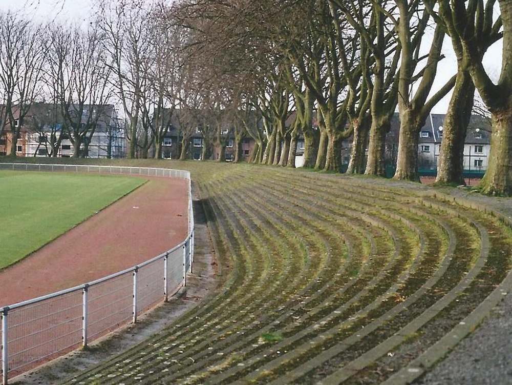 Stadion an der Bäuminghausstraße, TuS Helene Sicht auf eine geschwungene mit Gras zugewachsene Stehtribüne mit Bäumen im Hintergrund. Stadion an der Bäuminghausstraße, TuS Helene